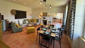 Un vaste espace de vie comprenant un canapé, une table basse et une table à manger, le tout prêt pour un repas. On y trouve également une cheminée, une cuisine à l’arrière-plan et une grande fenêtre avec des rideaux.