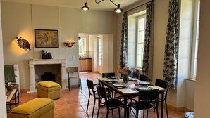 Un coin repas avec une longue table, des chaises et une cheminée. Il y a une fenêtre avec des rideaux et un tableau au mur.