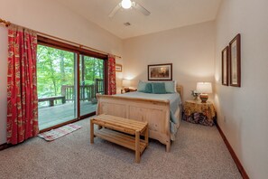 A bedroom with a wooden bed, a bench, a table with a lamp, and a view of a deck and trees. - New! Cozy Lakefront Retreat w/ Scenic Views (McHenry)