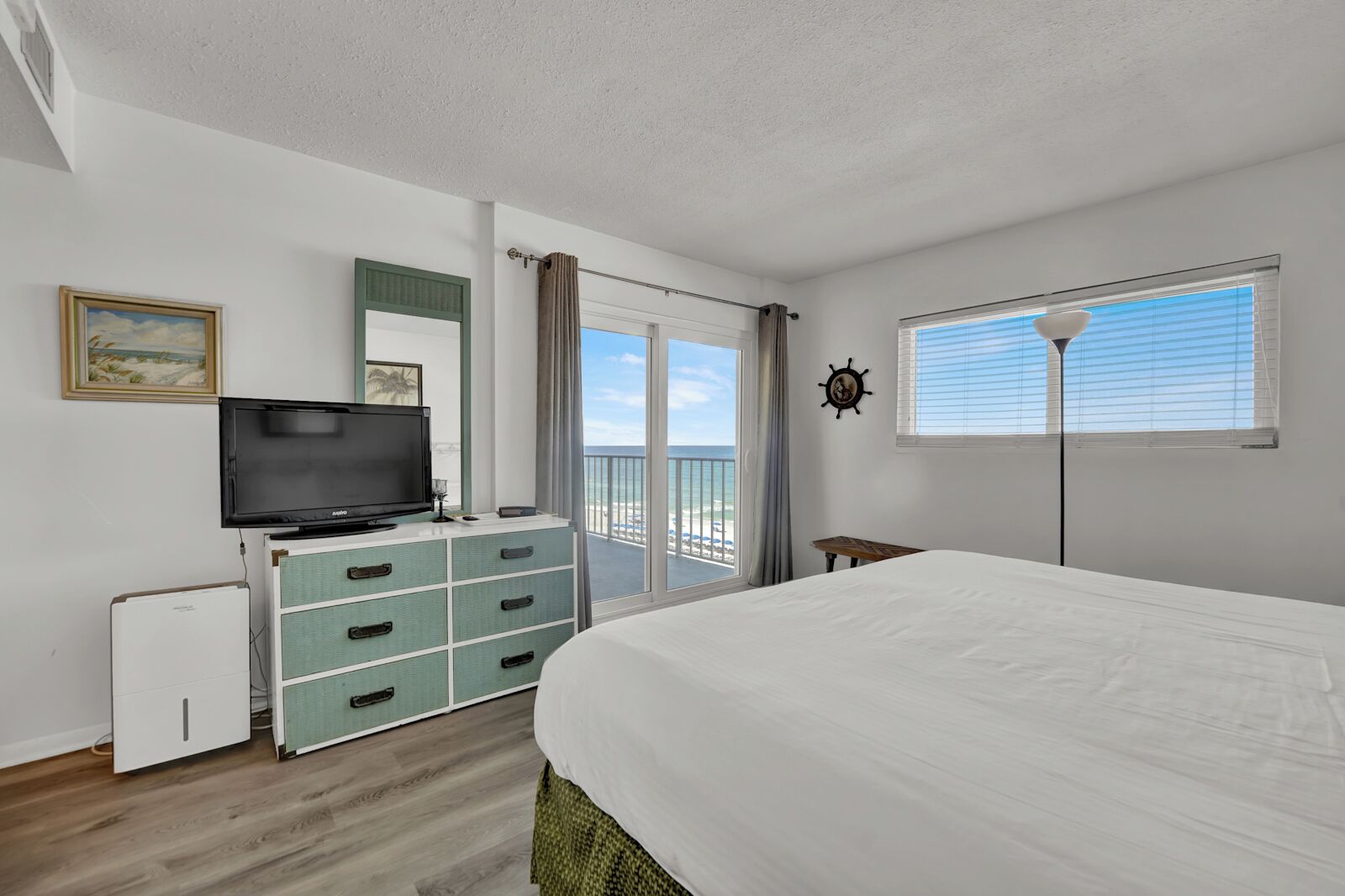 A hotel room with a bed, a television, a dresser, a small table, and a view of the ocean through the balcony doors.