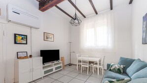 Una sala de estar con una televisión de pantalla plana sobre un mueble blanco, una mesa de comedor con sillas, un sofá azul y un ventanal con cortinas translúcidas.