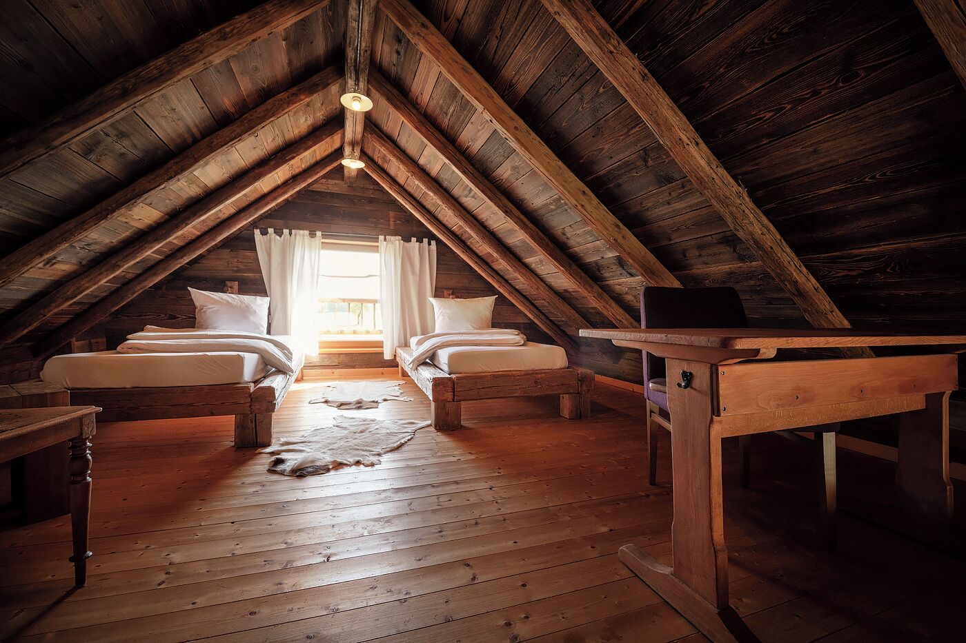 Quarto de sótão com duas camas, vigas de madeira, uma mesa e um candeeiro pendente.