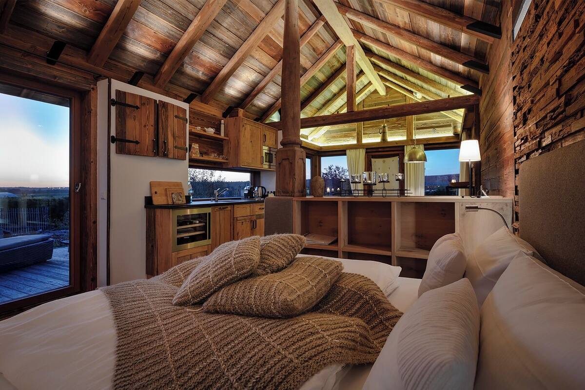 A cozy bedroom with a large bed, wooden beams, a view of the outdoors, a kitchenette, and a window with curtains.