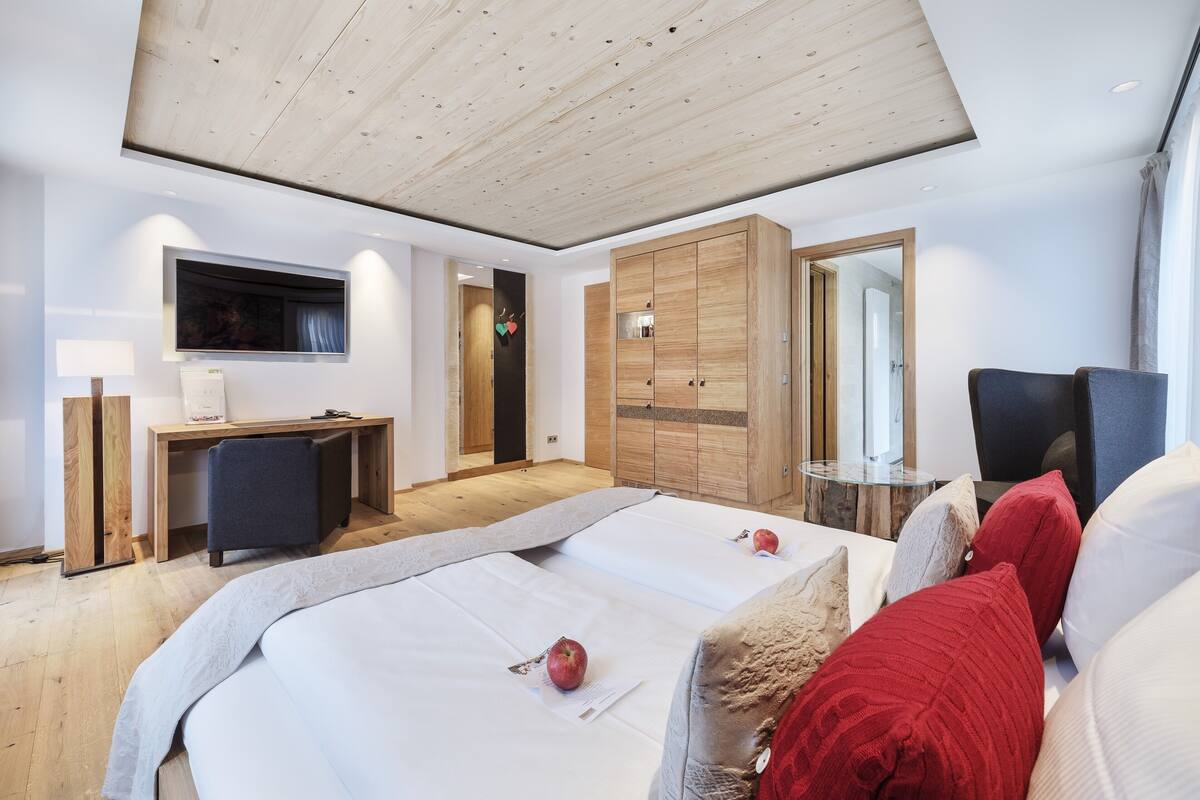 A modern hotel room with a wooden ceiling, a flat-screen TV mounted on the wall, a desk with a chair, a bed with white linens, and a large wardrobe.