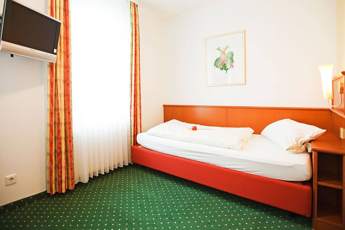 A hotel room with a single bed, a television mounted on the wall, and a painting above the bed.