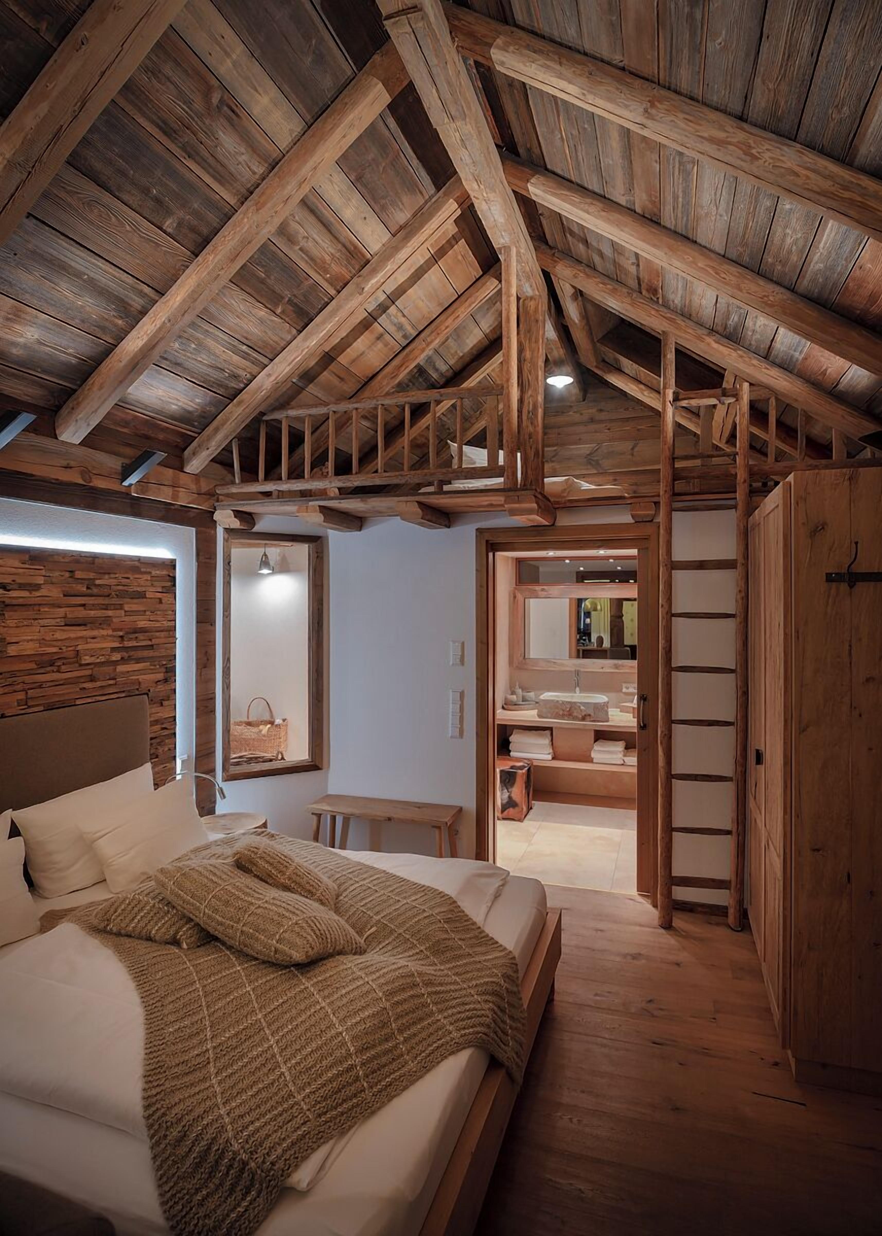 A rustic bedroom with a wooden ceiling, a bed with a knitted blanket, a wooden bench, and a bathroom visible through an open door.