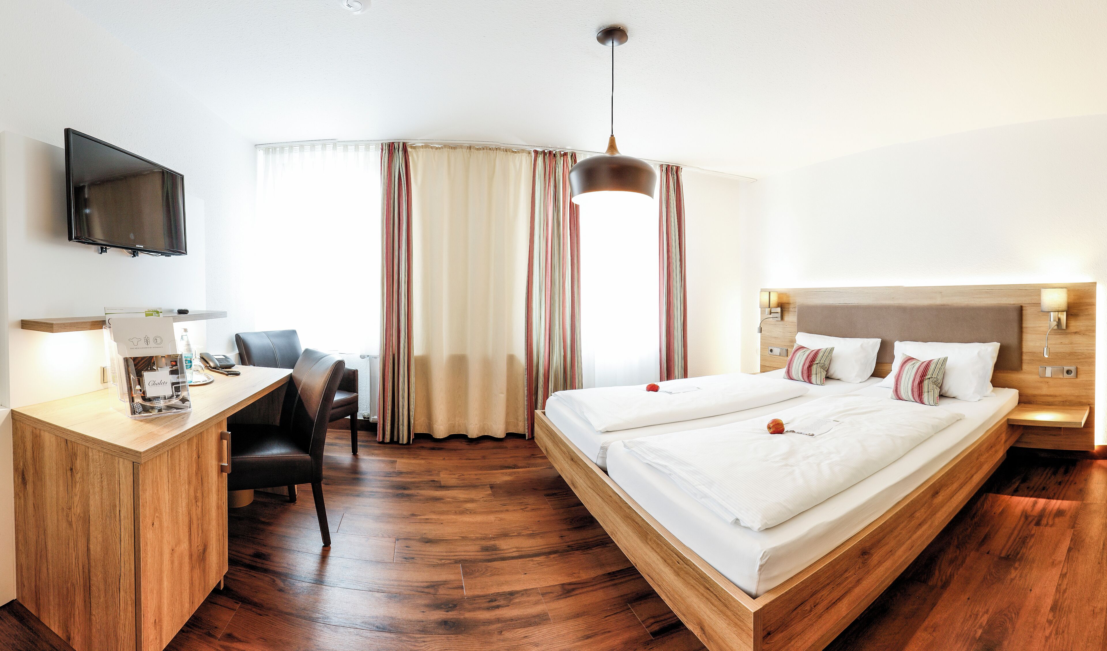 Une chambre d’hôtel avec un bureau en bois, un lit recouvert de draps blancs, un téléviseur fixé au mur et une fenêtre avec des rideaux.