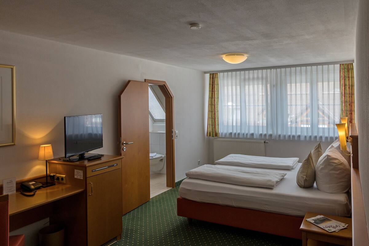 A hotel room with a bed, a desk with a lamp, a television, and a window with curtains.