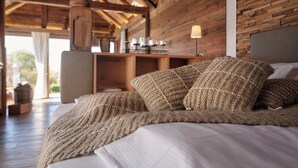 A bed with a knitted blanket and pillows, wooden beams, a lamp, and a view of the outdoors.
