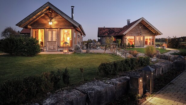 A wooden cabin with a large porch and a chimney, surrounded by a well-maintained garden and a stone wall.