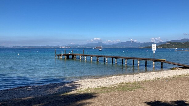 A wooden pier extending into calm water, with a rocky shoreline and distant mountains.