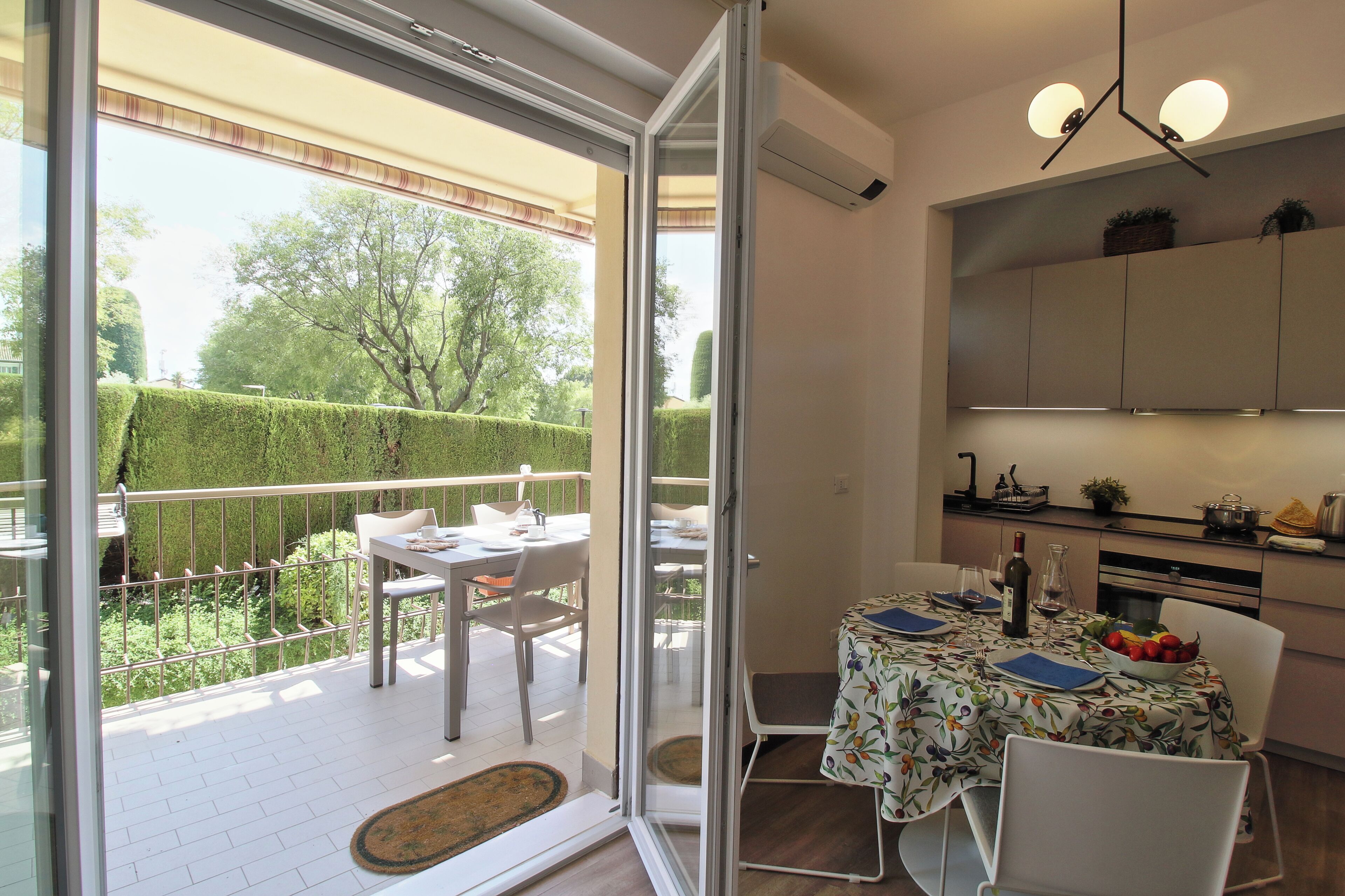 Een eetgedeelte met een tafel voor twee personen, een keuken met kasten en apparatuur, en uitzicht op een verzorgde tuin door een glazen deur.