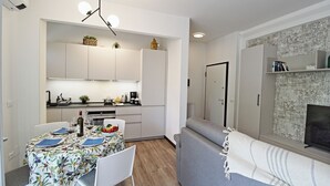 A modern kitchen with white cabinets, a dining table set for two, and a grey sofa in the living area.