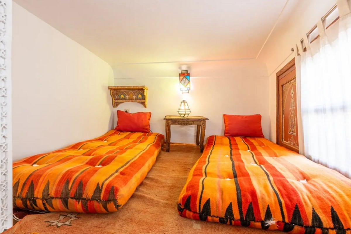 A room with two beds, a small table, a wall-mounted lamp, and a decorative wall piece.
