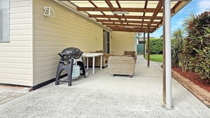 Een overdekt terras met een barbecue, tuinmeubilair en een uitzicht op een huis op de achtergrond.