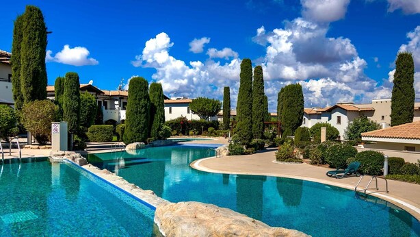 A swimming pool with a curved design, surrounded by neatly trimmed trees and a clear blue sky. - Junior Villa AA02 - Zeus (Kouklia)