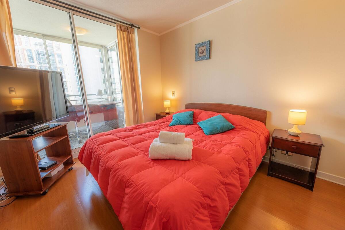 A hotel room with a large bed, a TV, a wooden side table, and a view through a sliding door.