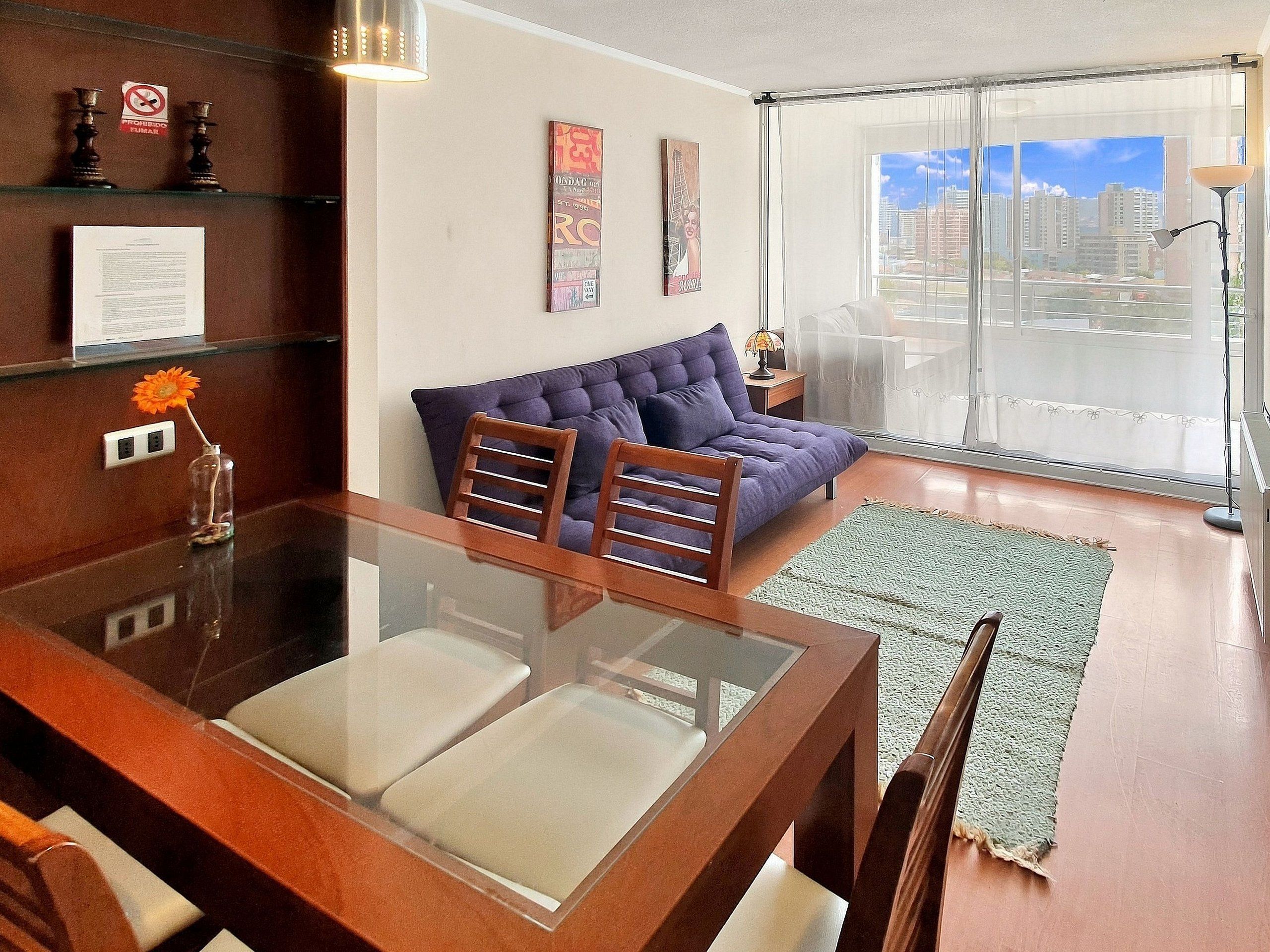 A modern living room with a glass dining table, a sofa, and a large window with a city view.
