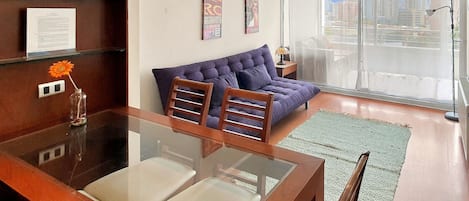 A modern living room with a glass dining table, a sofa, and a large window with a city view.