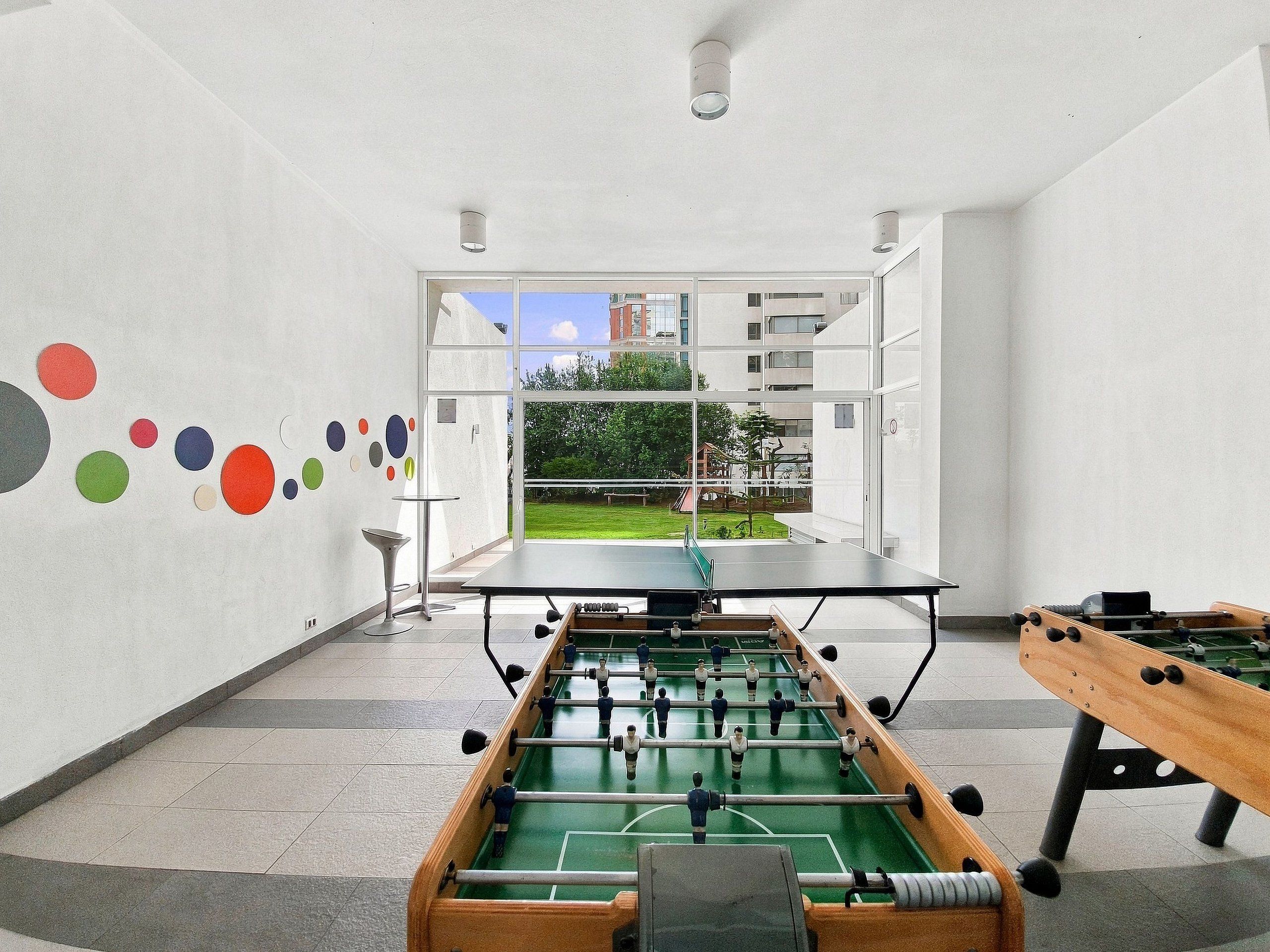 A room with a foosball table, a ping pong table, and a window with a view of a building and greenery.