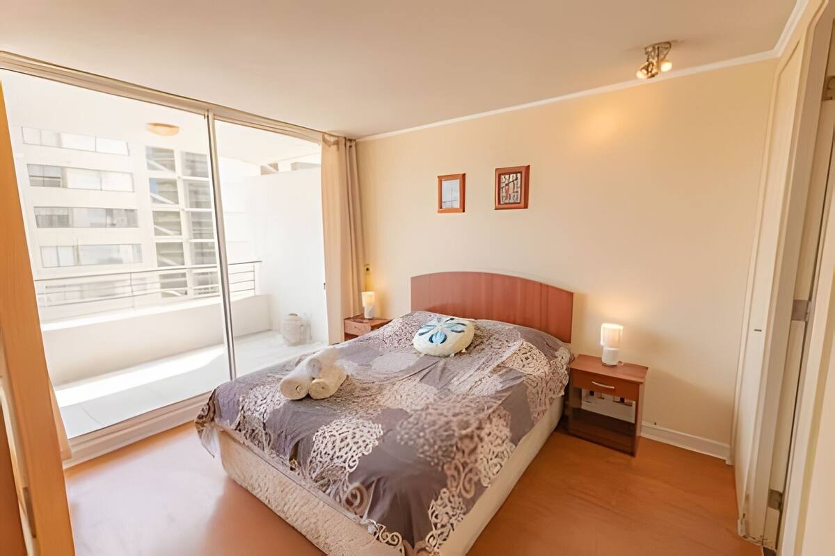 A bedroom with a large window, a bed with a patterned bedspread, two bedside tables with lamps, and a wall with two framed pictures.