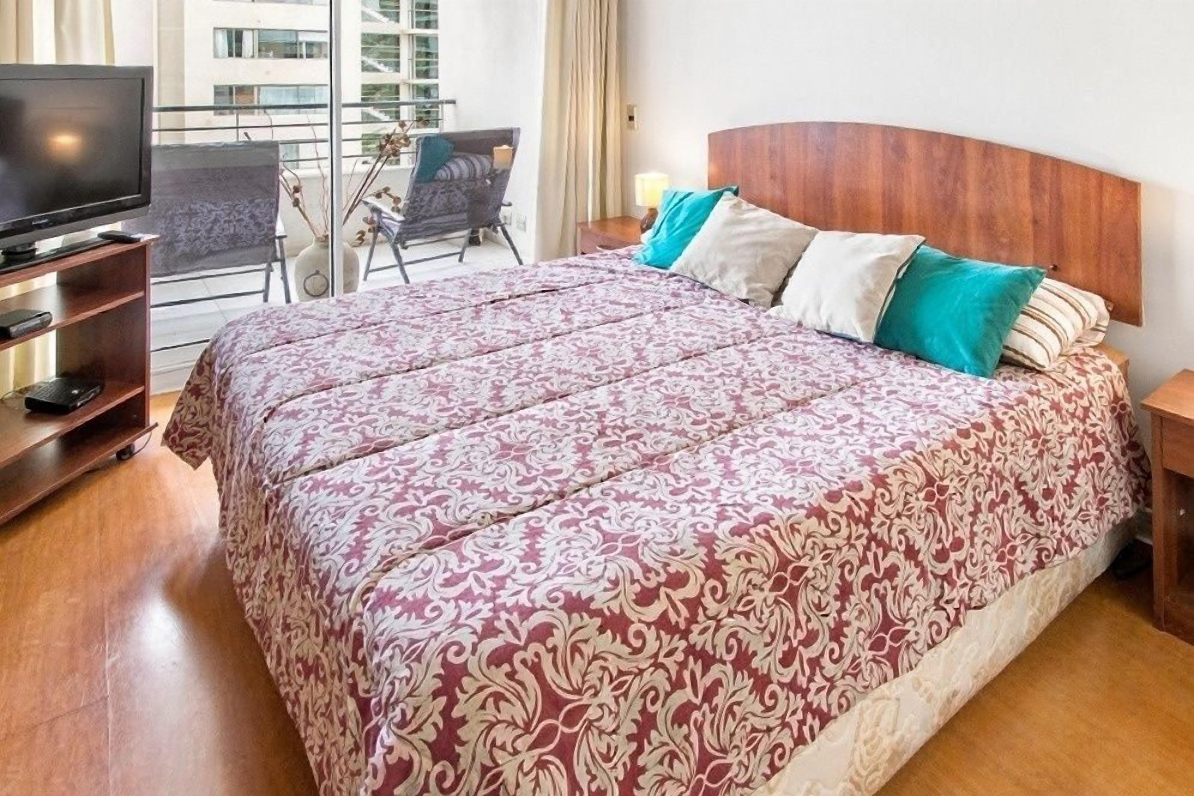 Habitación de hotel con una cama grande, un televisor sobre un mueble, un balcón con una silla y vistas a edificios.