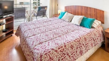 Habitación de hotel con una cama grande, un televisor sobre un mueble, un balcón con una silla y vistas a edificios.