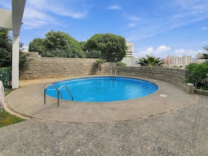 A circular swimming pool with a stone wall and metal ladder, surrounded by greenery and a clear sky.