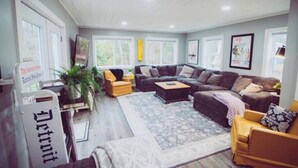 A spacious living room with a sectional sofa, a yellow armchair, and a coffee table. There are large windows providing natural light and a view of the outdoors. - RIVER FRONT Pickleball & Bball Patio-Pets-2 houses (Free Soil)