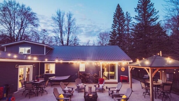 A covered outdoor patio with seating and a fire pit, surrounded by trees and a house. - RIVER FRONT Pickleball & Bball Patio-Pets-2 houses (Free Soil)