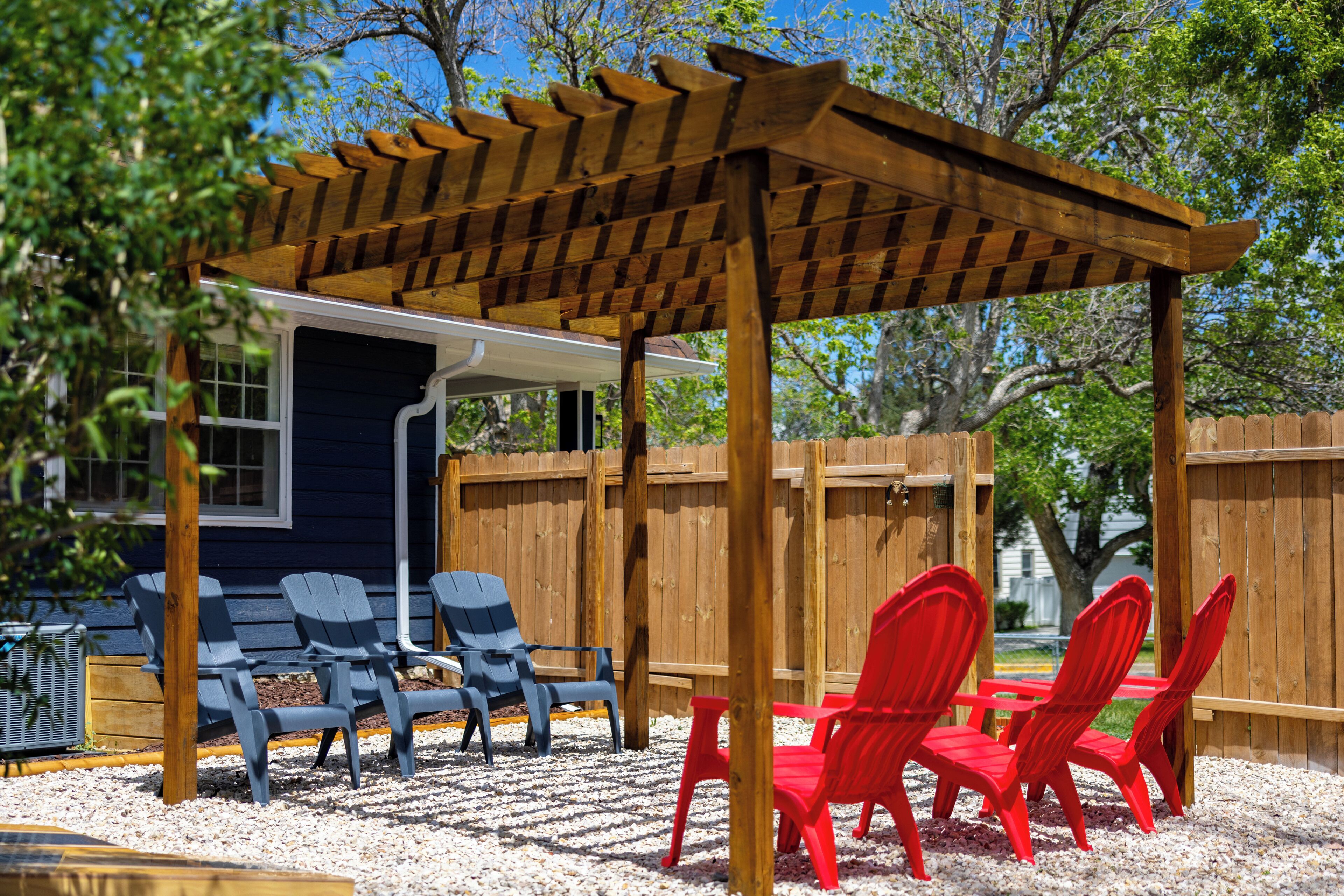 A covered outdoor seating area with wooden chairs and a table, surrounded by a wooden fence and trees.