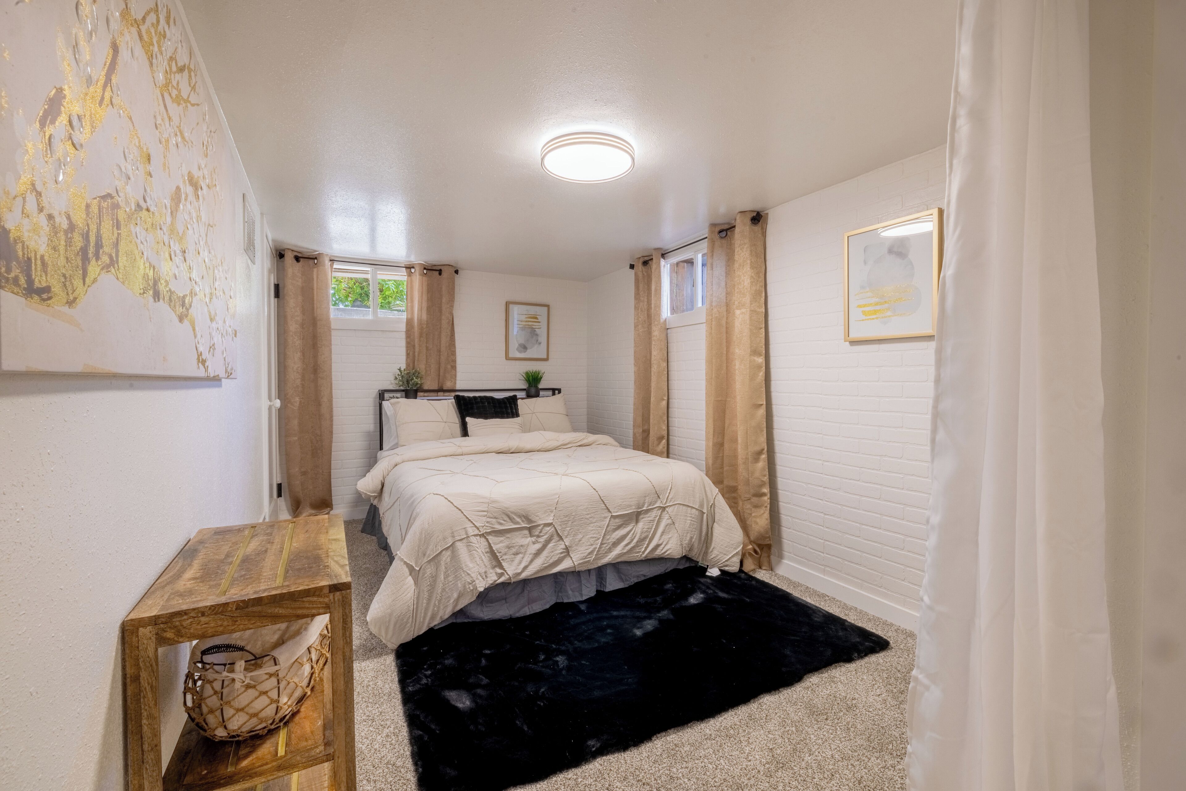 A bedroom with a bed, a small wooden table, a black rug, and a white wall with a framed picture.