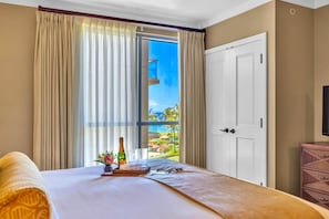 A hotel room with a bed, a small table with a bottle and glasses, and a view of the ocean through a window with curtains.