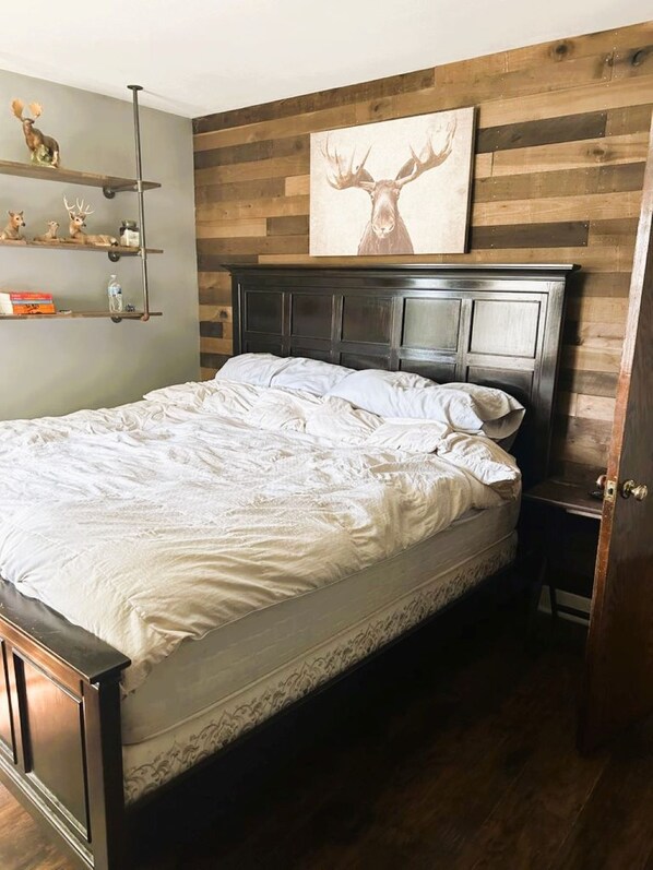 Quarto com cabeceira de madeira, cama com roupa de cama branca, prateleira com objetos decorativos e pintura de um veado.