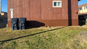 A small, single-story wooden building with a brown exterior, a single window, and two trash bins in front. - Clean & Quiet Studio Apartment B (Nome)