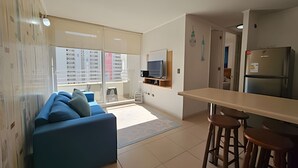 Ein modernes Wohnzimmer mit einem blauen Sofa, einer Küchenzeile inklusive Kühlschrank und einem Essbereich mit Hockern.