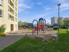 Un parque infantil con un tobogán rojo y estructuras para escalar, rodeado de césped y un edificio con balcones.