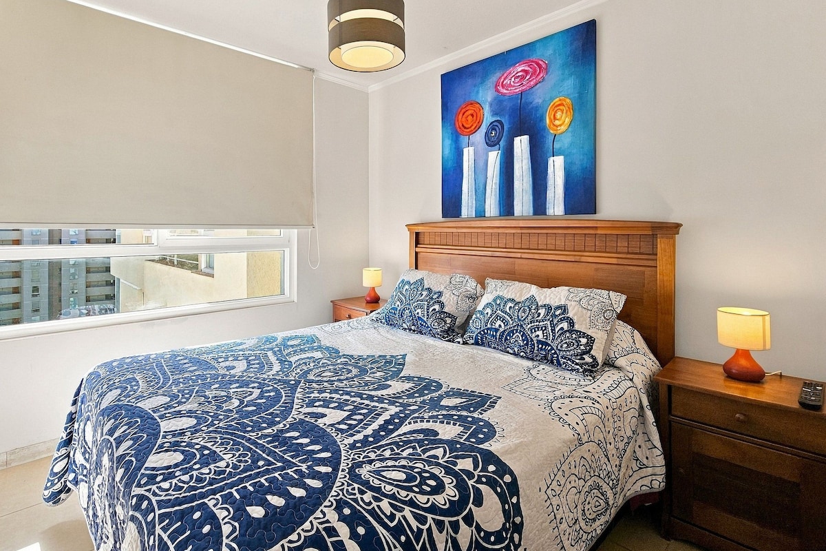 Une chambre à coucher avec un grand lit, une tête de lit en bois, un tableau au-dessus du lit et une fenêtre donnant sur des immeubles.