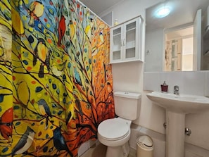 Une salle de bain avec un rideau de douche aux motifs colorés représentant des oiseaux, des toilettes blanches, un lavabo avec un miroir et un meuble de rangement.