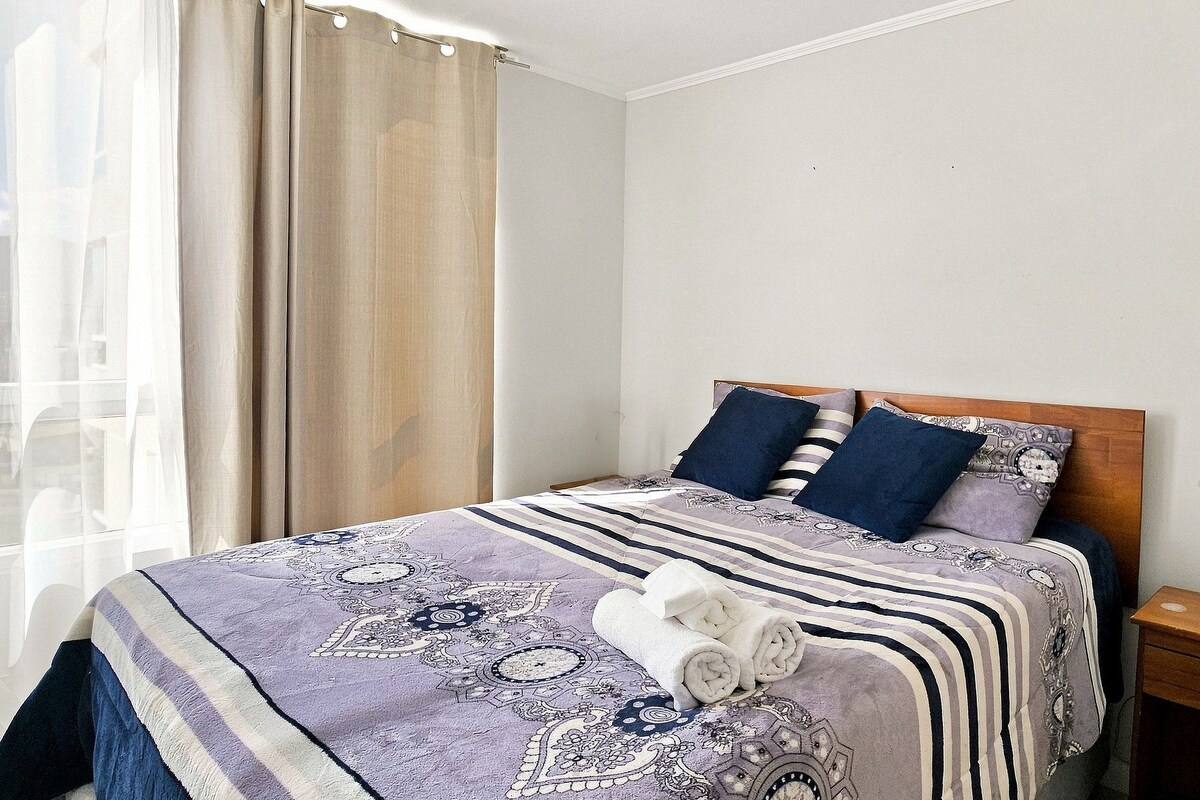 A hotel room with a bed, a nightstand, a lamp, and a window with curtains.