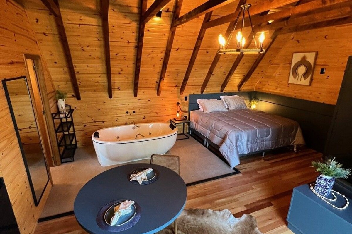 A cozy log cabin bedroom with a large bathtub, a bed, a dining table, and a chandelier.