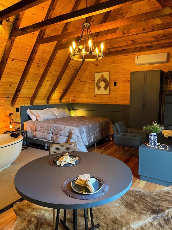 Una habitación acogedora y rústica con techo alto, una cama con cabecera, una mesa de comedor con posavasos y servilleta, una silla, un mueble bajo con una planta y un aire acondicionado de pared.