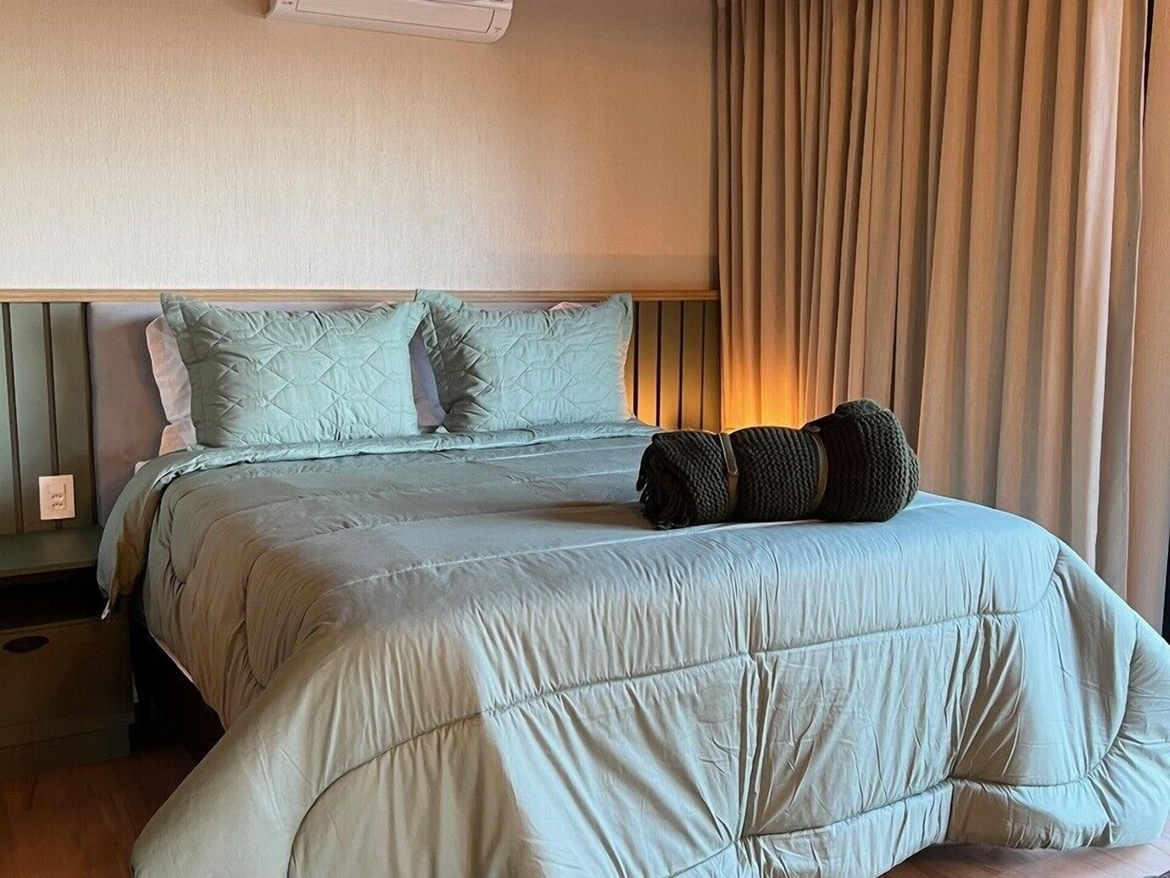Una cama bien hecha con un cabecero acolchado, una lámpara de noche y una ventana con cortinas.