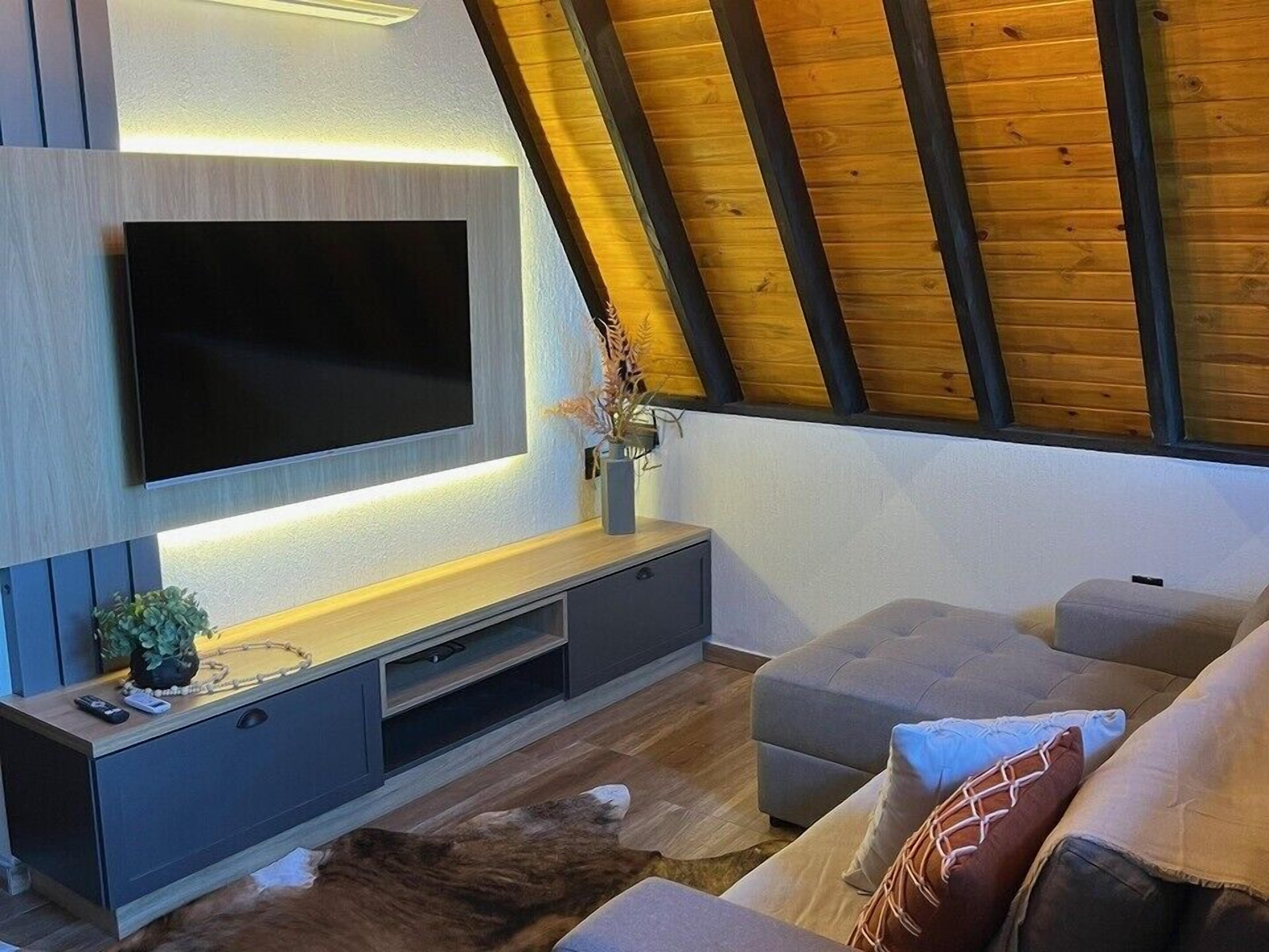 Una sala de estar moderna con un televisor de pantalla plana montado en una pared de madera, un sofá gris y un techo con vigas a la vista.