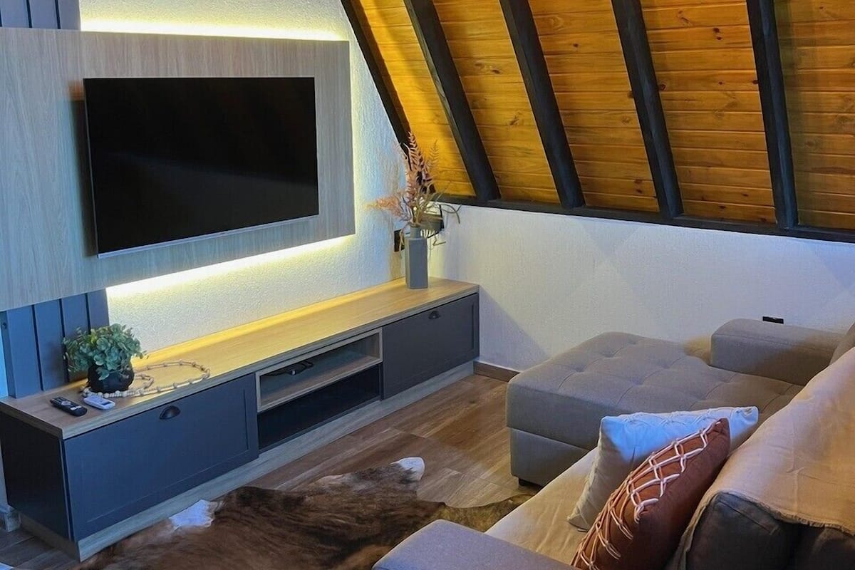 A modern living room with a flat-screen TV mounted on a wooden wall, grey sofa, and a ceiling with exposed beams.