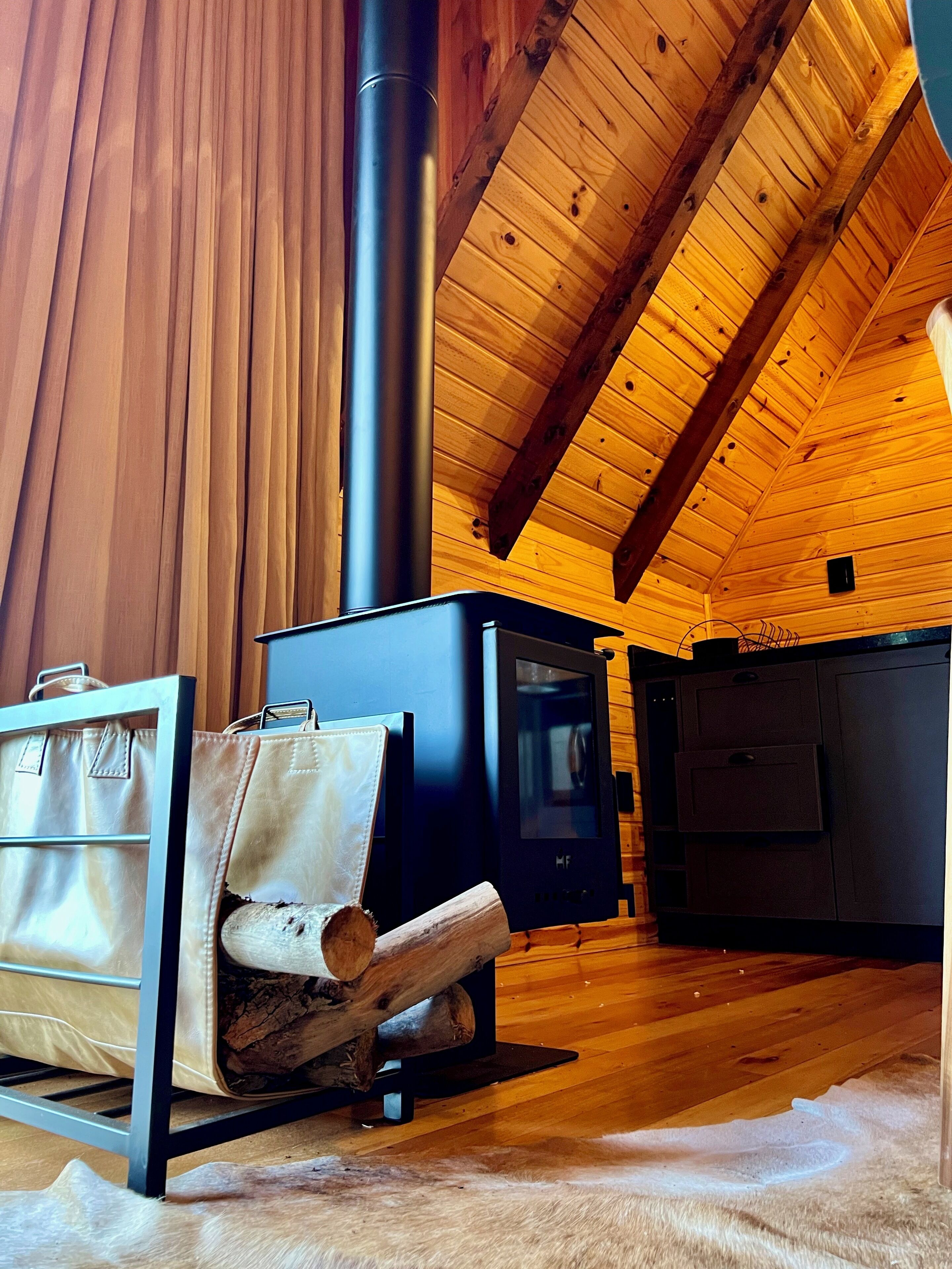 Interior acogedor de una cabaña con estufa de leña, un sofá y un bolso, y techo de madera.