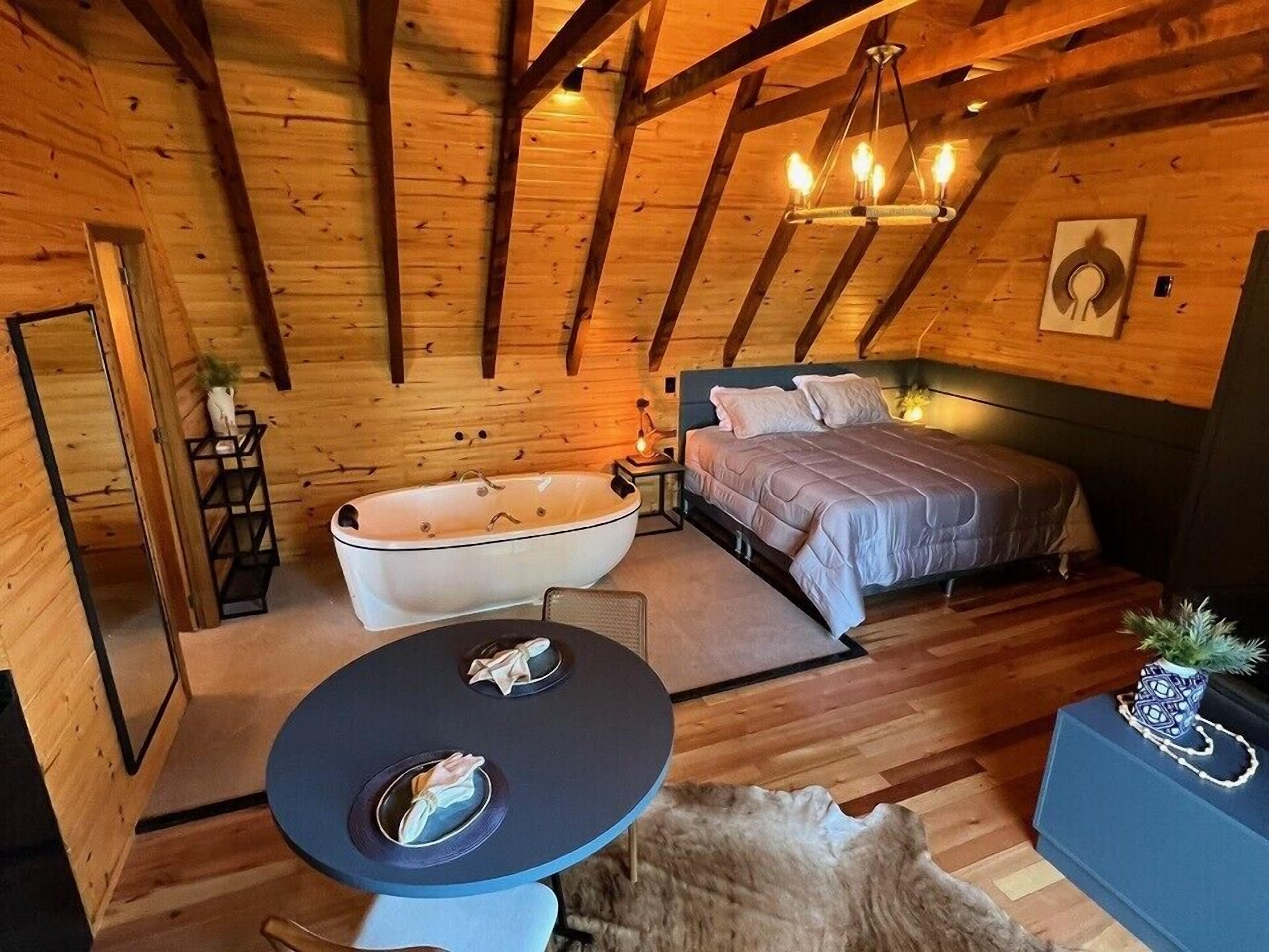 Acogedor dormitorio de cabaña de madera con una bañera grande, una cama, una mesa de comedor y una araña.