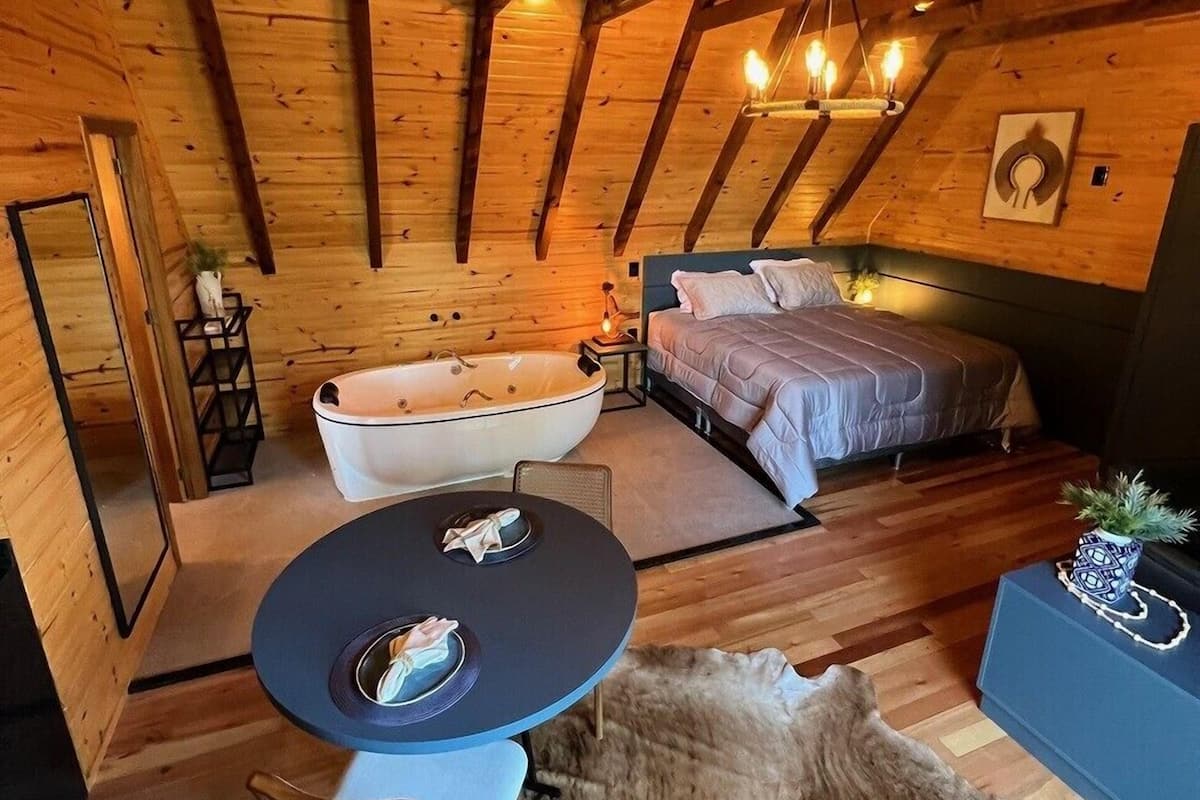 A cozy log cabin bedroom with a large bathtub, a bed, a dining table, and a chandelier.