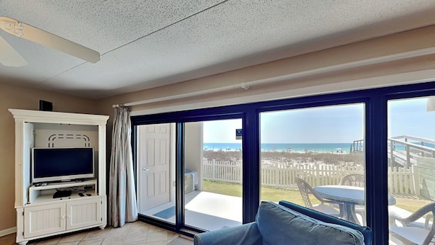 A living room with a TV, a sofa, and a balcony with a view of the beach. - Ocean Breeze West #106 | Gulf-Front, Ground Floor, Pool, Beach Access (Perdido Key)
