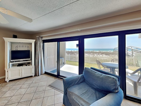 A living room with a TV, a sofa, and a balcony with a view of the beach.